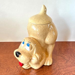 May include: A ceramic cookie jar shaped like a tan dog in a playful pose with its rear end raised. The dog has a glossy finish, bright blue eyes, a black nose, and a small red tongue sticking out. The top portion of the dog's back forms a lid.