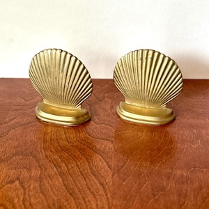 May include: Two gold-colored seashell bookends. Each bookend has a fan-shaped design with vertical lines, mounted on a rectangular base. The bookends are displayed on a wooden surface.