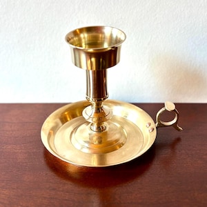 Peut inclure: Un bougeoir en laiton doré avec une base circulaire, une tige centrale et un sommet en forme de coupe. Il possède une petite poignée et est conçu pour une seule bougie. Le laiton a une teinte dorée. Hauteur d'environ 15 cm.