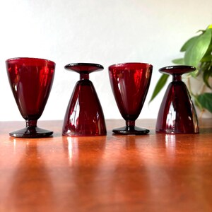 May include: Four deep red glass goblets, two standing upright on circular bases and two inverted, showcasing a smooth, translucent finish. The stemware features a classic tapered bowl shape and a consistent dark ruby hue throughout the glass.