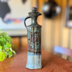 May include: A tall, ceramic olive oil bottle with a handle and spout. The bottle is primarily brown and blue with floral designs and a light gray base. The lid has a small, decorative knob. The bottle is on a wooden surface.