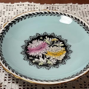 May include: A small, round, light blue dish with a gold rim and scalloped edge. The center features a black, ornate design with pink and yellow floral details. A black and white leaf pattern borders the rim.