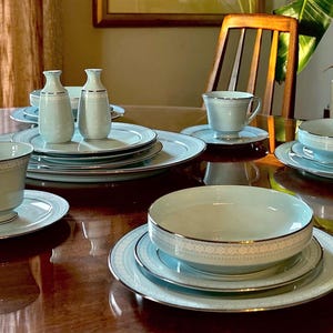 Op de afbeelding: Een compleet lichtblauw en wit porseleinen servies met zilveren rand. De set bevat borden, kommen, kopjes, schotels en kleine decoratieve flesjes. Het servies staat op een reflecterende houten tafel.