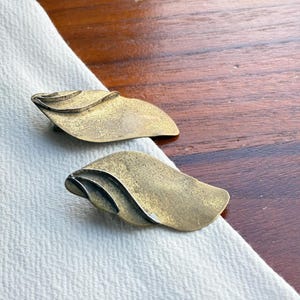 Op de afbeelding: Twee vintage oorbellen in de vorm van een blad, in messingkleur. Elke oorbel heeft een gelaagd ontwerp met een getextureerd oppervlak. De oorbellen worden gepresenteerd op witte stof, tegen een donkere houten achtergrond.