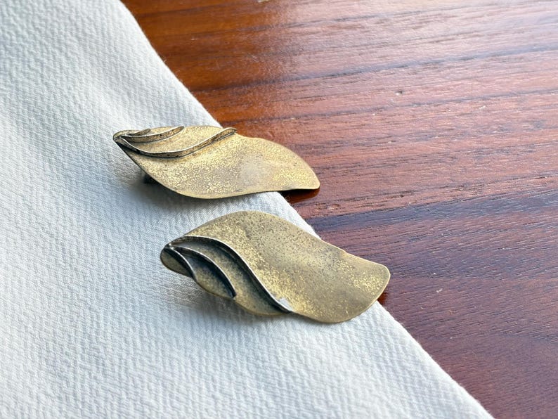 Op de afbeelding: Twee vintage oorbellen in de vorm van een blad, in messingkleur. Elke oorbel heeft een gelaagd ontwerp met een getextureerd oppervlak. De oorbellen worden gepresenteerd op witte stof, tegen een donkere houten achtergrond.