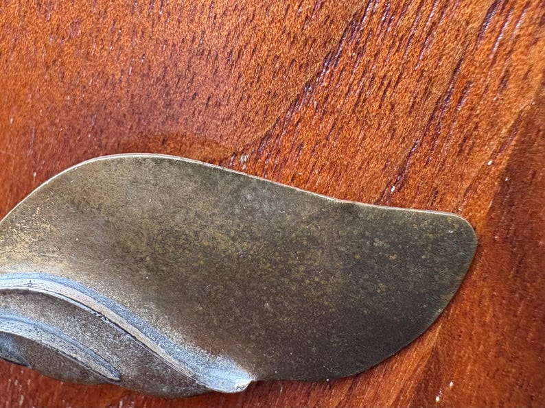 Op de afbeelding: Close-up van een metalen, bladvormig object met een getextureerd oppervlak. Het object heeft een donkere, verouderde uitstraling, met hints van groen en bruin. De achtergrond is een rijke, roodbruine houtnerf.