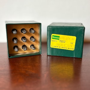 Könnte beinhalten: Ein Satz Metall-Zahlenstempel in einer grünen Box mit Holzeinsatz. Der Deckel trägt ein gelbes Etikett mit dem Text HANSON STD. DTY. NO. 386313 1/8 und H.L. HANSON, INC., WORCESTER, MA 01606. Die Stempel sind in einem Raster angeordnet.
