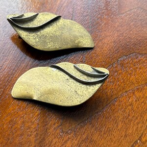 Op de afbeelding: Twee vintage decoratieve objecten in de vorm van bladeren van messing. Elk heeft een getextureerd, gouden oppervlak met een zilverkleurig, golvend accent. De objecten worden gepresenteerd op een donker houten oppervlak, wat hun unieke ontwerp laat zien.