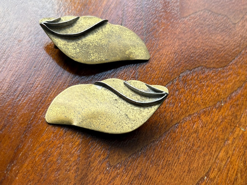 Op de afbeelding: Twee vintage decoratieve objecten in de vorm van bladeren van messing. Elk heeft een getextureerd, gouden oppervlak met een zilverkleurig, golvend accent. De objecten worden gepresenteerd op een donker houten oppervlak, wat hun unieke ontwerp laat zien.