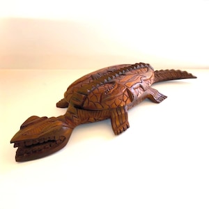 May include: A handcrafted wooden alligator sculpture with intricate carvings. The alligator is brown with a detailed textured surface, including scales and teeth. The sculpture has a long body and tail, and is lying down.
