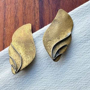 Op de afbeelding: Een paar oorbellen in vintage stijl. Elke oorbel heeft een bladachtige vorm met een getextureerde, goud-bronzen afwerking. Het ontwerp heeft gelaagde elementen en een zilverkleurige accentlijn, tegen een witte stoffen achtergrond.