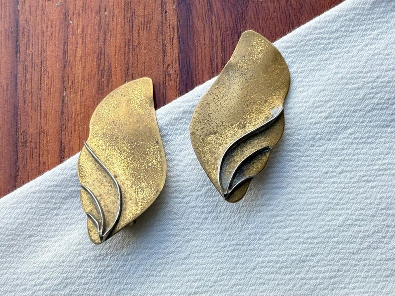 Op de afbeelding: Een paar oorbellen in vintage stijl. Elke oorbel heeft een bladachtige vorm met een getextureerde, goud-bronzen afwerking. Het ontwerp heeft gelaagde elementen en een zilverkleurige accentlijn, tegen een witte stoffen achtergrond.