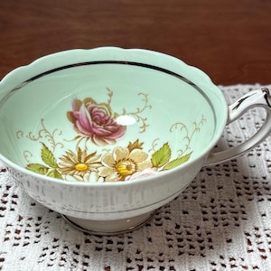 Puede incluir: Una taza de té vintage con un interior verde claro y un diseño floral con una rosa rosa y margaritas amarillas. La taza tiene un exterior blanco con un borde y asa plateados. La taza de té está sobre un tapete de encaje blanco.