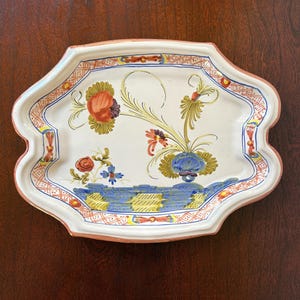 May include: An oval ceramic serving platter with a scalloped edge and a pink rim. The center features hand-painted floral motifs in blue, orange, yellow, and green, surrounded by a decorative orange and white lattice border pattern on the rim.
