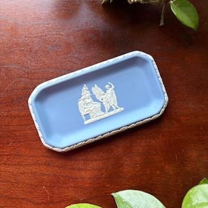 May include: A rectangular light blue jasperware tray featuring a white relief scene of three figures and a decorative white border. The central relief depicts a seated figure, a winged figure, and a standing figure in a classical style.