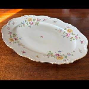 May include: An oval, white porcelain serving platter with a scalloped edge and gold trim. The platter is decorated with floral patterns in yellow, pink, blue, and green. A classic piece for serving food.