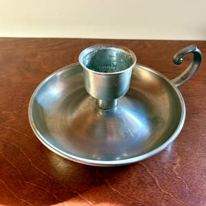 May include: A silver-toned metal candlestick holder with a circular base and a central cup for holding a candle. It features a curved handle on the side. The candlestick holder is displayed on a wooden surface.