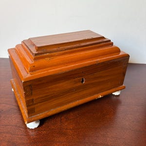 May include: A rectangular wooden box with a polished medium-brown finish, featuring a stepped lid design, visible dovetail joinery on the corners, a central keyhole on the front panel, and four small, white, scalloped-shaped feet at the base.