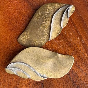 Op de afbeelding: Twee abstracte, bladvormige metalen objecten met een goud- en zilverafwerking. De bladeren hebben een textuur en zijn gerangschikt op een houten achtergrond. Het ontwerp heeft gelaagde, gebogen elementen.