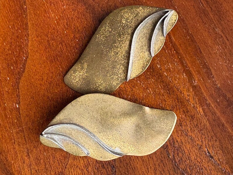 Op de afbeelding: Twee abstracte, bladvormige metalen objecten met een goud- en zilverafwerking. De bladeren hebben een textuur en zijn gerangschikt op een houten achtergrond. Het ontwerp heeft gelaagde, gebogen elementen.