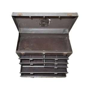 Puede incluir: Caja de herramientas metálica gris, abierta, con tapa con bisagras y varios cajones extraíbles. La caja muestra signos de desgaste, lo que sugiere antigüedad y uso. El interior es oscuro.