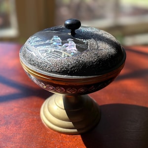 May include: A black and gold decorative pedestal bowl with a lid. The lid features a mother-of-pearl inlay scene of figures on a bridge. The bowl has a gold band and sits on a brass-colored pedestal base.