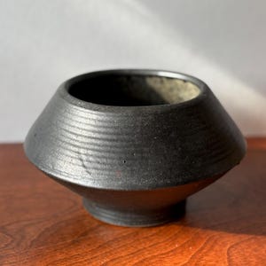 Peut inclure: Un bol en céramique noir mat avec une épaule large et inclinée et une base étroite. La surface extérieure présente de subtiles textures striées horizontales entourant la partie supérieure, tandis que l'intérieur révèle une finition mouchetée plus claire.