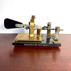 May include: A vintage telegraph sounder featuring a black base with the text SOUNDER 3C 140 OHMS printed in white. The device includes a brass arch, a metal lever with a black handle, and various adjustment screws and metal components.