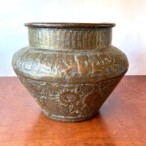 Può includere: Un vaso in ottone a bocca larga con intricate incisioni fatte a mano. La fascia superiore presenta un motivo verticale simile a una recinzione, mentre il corpo mostra dettagliate rappresentazioni in rilievo di un sole, motivi floreali e figure umane.