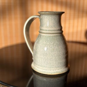 Puede incluir: Una jarra de cerámica hecha a mano con asa. La jarra es de color gris verdoso claro con una textura moteada. Tiene una base ancha que se estrecha hacia el cuello y un borde acampanado. El asa es curva y está unida al lateral de la jarra.