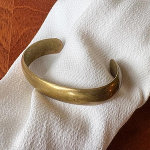 May include: A gold-colored cuff bracelet with a curved design. The bracelet is resting on a white, textured fabric. The background includes a wooden surface.
