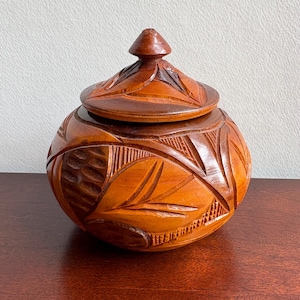 May include: A round, medium-brown wooden lidded jar featuring intricate hand-carved patterns across its surface. The design includes stylized leaf motifs, cross-hatched textures, and smooth, curved lines. The lid has a pointed, conical handle.