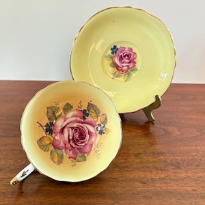 Puede incluir: Un juego de taza y platillo de té vintage. La taza y el platillo son de color amarillo pálido con un diseño floral de rosas en tonos rosas y verdes. La taza tiene un asa blanca con ribete dorado.