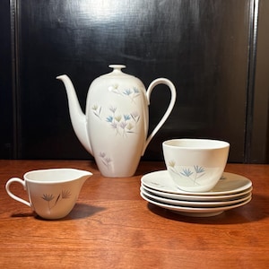 May include: A white ceramic tea set with a teapot, creamer, cup, and saucers. The set features a delicate floral design in blue, green, and purple. The teapot has a curved spout and handle. The cup and saucers are stacked.
