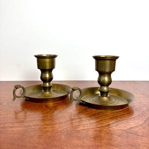Puede incluir: Un par de candelabros de latón a juego, cada uno con una base circular ancha, un pedestal central con la parte superior acampanada y una pequeña asa curva unida al lateral de la base. El metal tiene un acabado pulido de color marrón dorado.