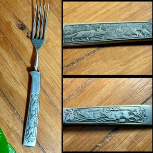 Puede incluir: Un tenedor plateado vintage con un mango detallado. El mango presenta escenas de fauna grabadas, incluyendo un ciervo y un zorro. El tenedor tiene púas delgadas y un mango rectangular con bordes redondeados. Las palabras "STAINLESS GERMANY" están estampadas en el tenedor.