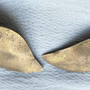 Op de afbeelding: Close-up van twee goudkleurige oorbellen in de vorm van bladeren. De oorbellen hebben een textuur en een lichte curve, wat wijst op een natuurlijk ontwerp. De achtergrond is een lichtgekleurde, getextureerde stof.