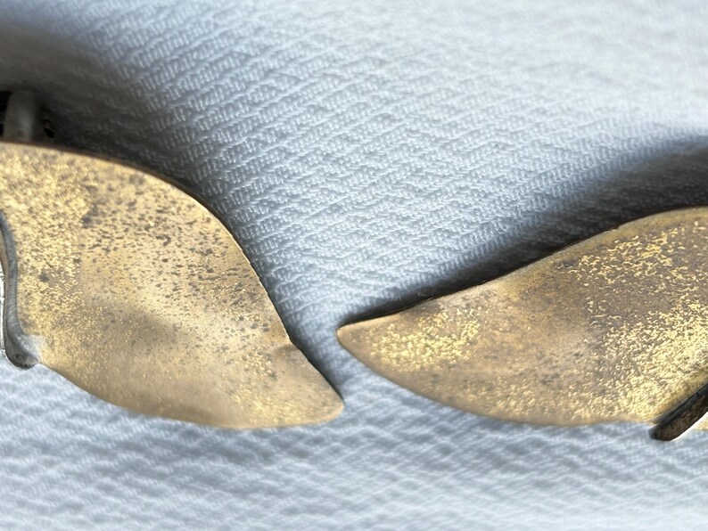 Op de afbeelding: Close-up van twee goudkleurige oorbellen in de vorm van bladeren. De oorbellen hebben een textuur en een lichte curve, wat wijst op een natuurlijk ontwerp. De achtergrond is een lichtgekleurde, getextureerde stof.