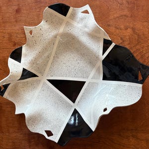 May include: A decorative ceramic bowl with a unique, geometric design. The bowl features a speckled white interior with black and white geometric shapes and a scalloped edge. The bowl has a modern, artistic aesthetic.