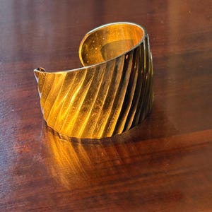 May include: A wide, gold-colored cuff bracelet with a textured, ridged design. The bracelet has a curved shape and reflects light, creating highlights and shadows. The jewelry is made of metal and appears to be a statement piece.