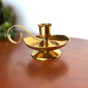 May include: A polished gold candle holder with a curved handle. The holder features a circular base and a small cup for a candle. The design is simple and elegant, suitable for various decor styles. The candle holder is approximately 10 cm in diameter.