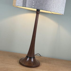 May include: A table lamp with a dark brown wood grain base that tapers upwards to a thin neck. The lamp features a cylindrical fabric lampshade with a textured grey, black and tan striped pattern. The light is illuminated from within the shade.