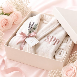 May include: A cream-colored plush robe with the embroidered initials "SPP" in pink, presented in a gift box. The robe is tied with a pink ribbon and a tag that reads "With Love". The box is surrounded by pink roses and baby's breath.