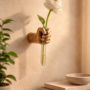 May include: A gold-coloured hand-shaped wall vase holding a white flower with a green stem and leaves. The vase is a clear glass tube. The wall is a light beige colour. A small plant and books are on a wooden surface.