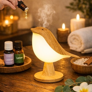 Może przedstawiać: Drewniany dyfuzor olejków eterycznych w kształcie ptaka z jasno białym korpusem i brązowymi drewnianymi elementami. Dyfuzor stoi na drewnianym stole, w tle butelki z olejkami eterycznymi i świece. Ręka wlewa olej.