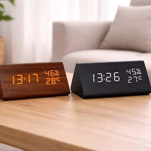May include: Two triangular digital clocks on a wooden table. One is dark wood with orange display, showing time, humidity, and temperature in Celsius. The other is black with a white display, showing time, humidity, and temperature in Celsius.