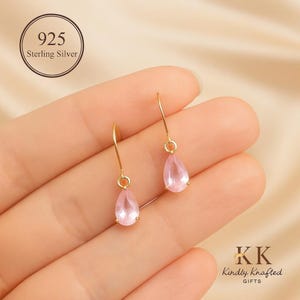 May include: A pair of gold-coloured drop earrings featuring teardrop-shaped, light pink gemstones. The earrings hang from delicate hooks and are held in a hand. The earrings are labelled "925 Sterling Silver".