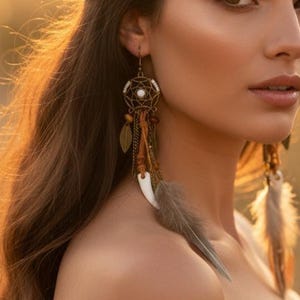 May include: A pair of dangling earrings featuring a circular dreamcatcher frame in an antique bronze finish, accented with a central white bead, brown leather cords, small metallic leaf charms, and long, soft grey feathers hanging from the base.