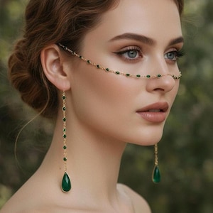 May include: A gold-coloured face chain and earrings set. The face chain is draped across the nose and forehead, decorated with small green beads. The earrings have a long chain with green beads, ending in a teardrop-shaped green gem.