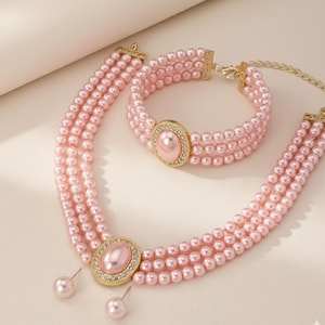 May include: A pink pearl necklace, bracelet, and earring set. The necklace and bracelet feature multiple strands of small, round pearls. Each piece has a gold-colored oval centerpiece with a pink pearl and surrounding clear crystals. The earrings are pink pearl studs.